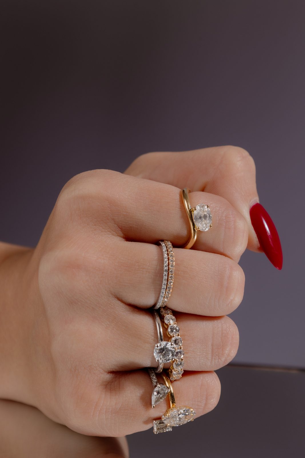 Poing fermé portant des bagues en or et diamants empilées, ongles rouges, sur fond gris